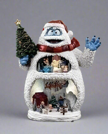 Animated snowman with a small village scene inside, waving and holding a Christmas tree on a black background.